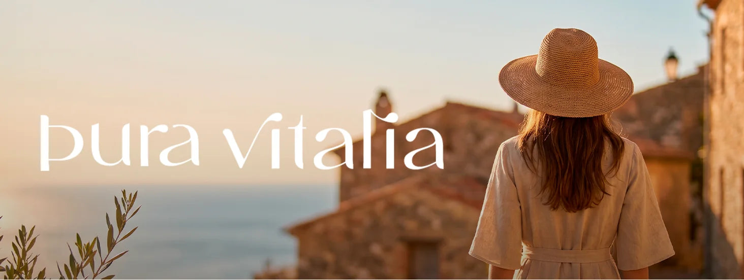 A woman in a straw hat looking out over a Mediterranean village at sunset, with the Pura Vitalia wordmark